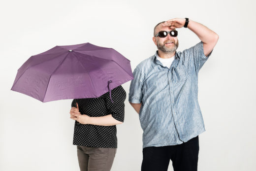 Kaksi henkilöä, toinen piiloutunut sateenvarjon alle, toinen hymyilee ja tähyilee aurinkolasit silmillä.
